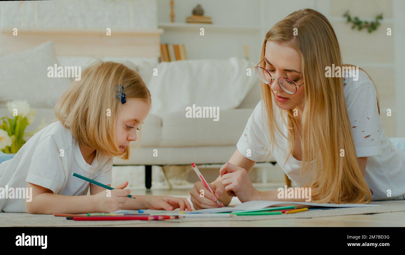 Piccola bambina adottiva godere di disegno d'arte con la madre caucasica in occhiali che giacciono a casa piano insieme. Bambino figlia con mamma mommia Foto Stock