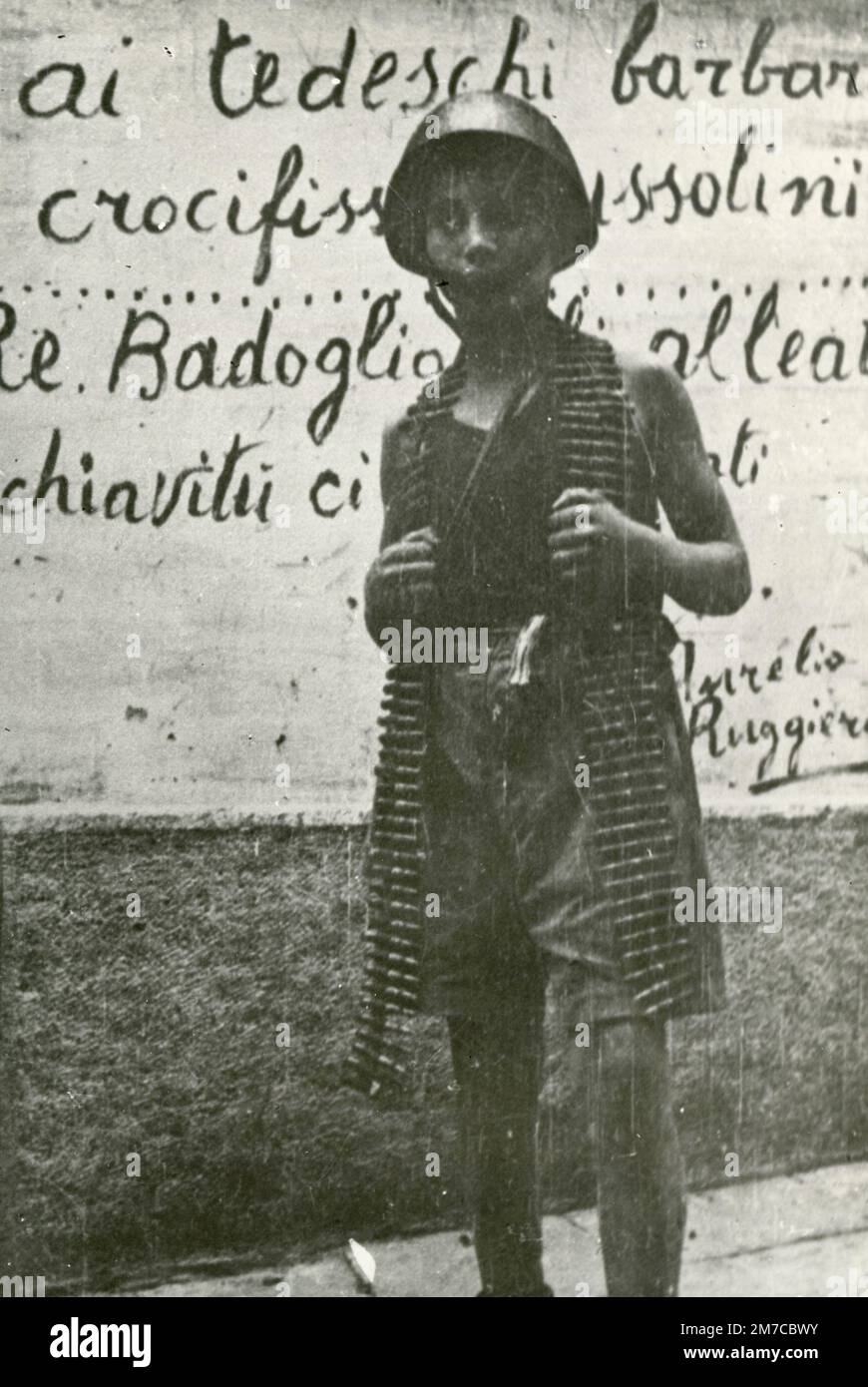Bambino con casco e munizioni vicino ad un scritto sulla parete dopo il WW2, Italia 1940s Foto Stock