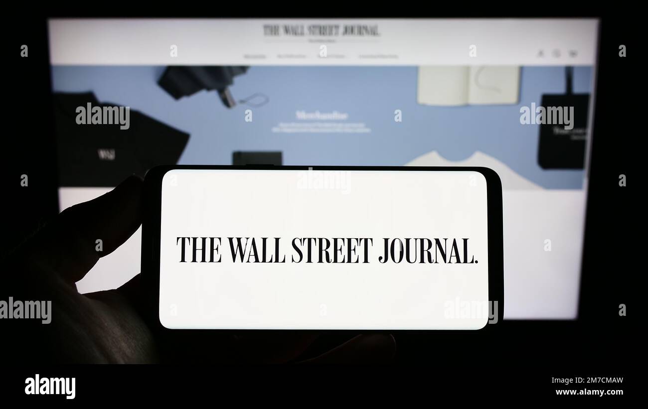 Persona che tiene in mano uno smartphone con il logo del quotidiano statunitense The Wall Street Journal sullo schermo di fronte al sito Web. Messa a fuoco sul display del telefono. Foto Stock