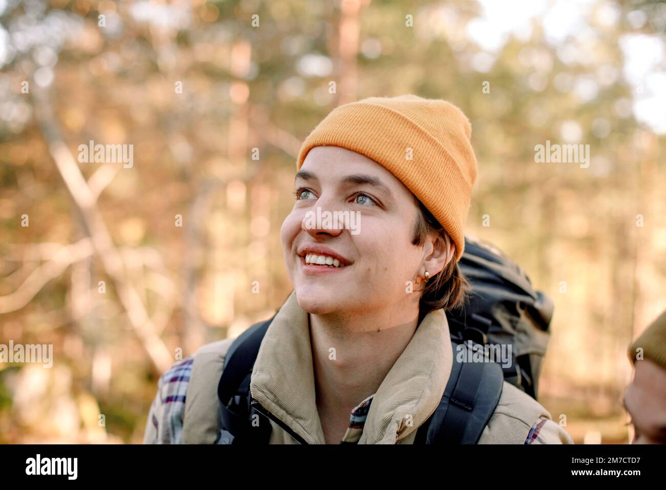 Giovane uomo sorridente che indossa un cappello a maglia che guarda lontano durante le escursioni Foto Stock