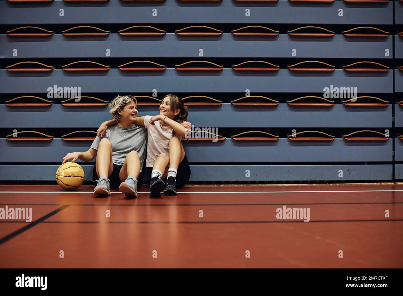 Tutta la lunghezza delle ragazze sorridenti che si siedono con le braccia intorno a un campo di pallacanestro a scuola Foto Stock