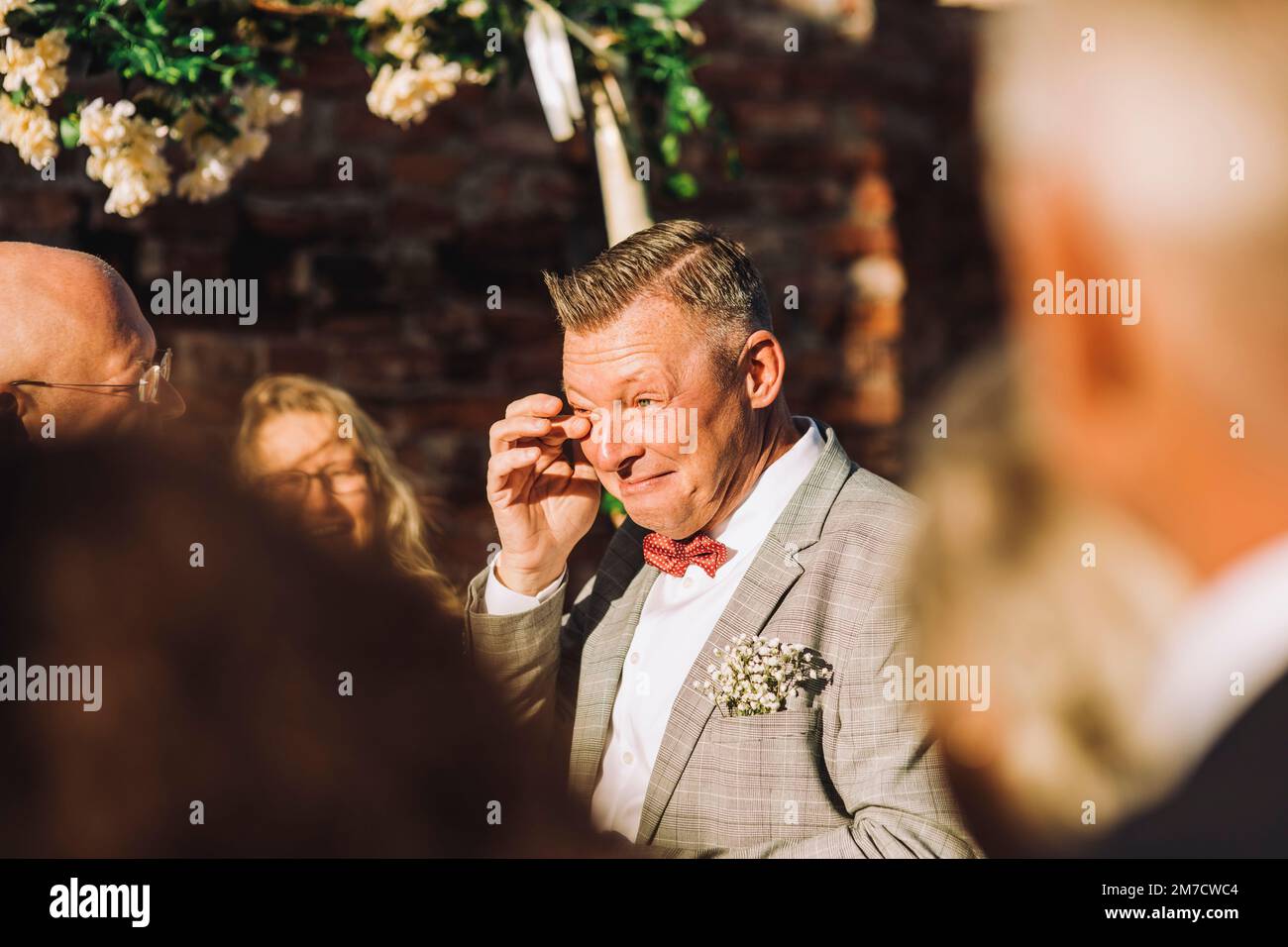 Sposo sorridente strofinando le sue lacrime con il dito in mezzo agli ospiti nella giornata di sole Foto Stock