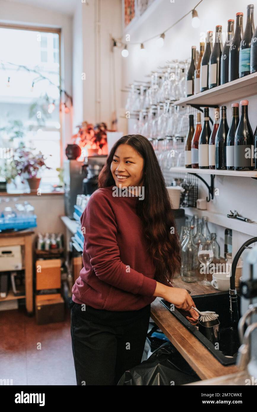 Donna sorridente proprietaria di una caffetteria che lavava piatti nella caffetteria Foto Stock