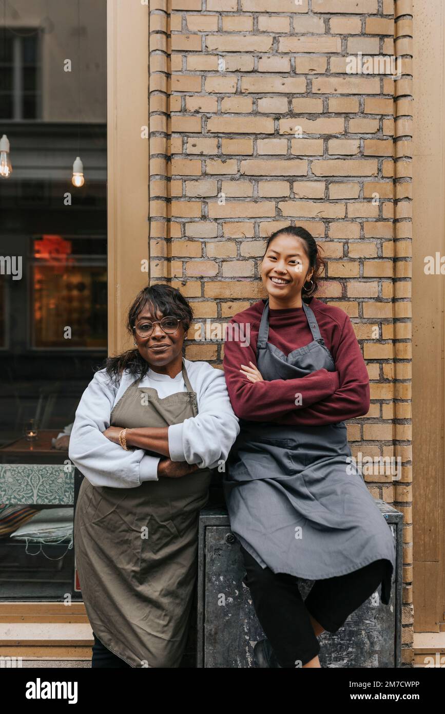 Ritratto delle donne sorridenti proprietari di caffè che indossano grembiuli in piedi con le braccia incrociate all'esterno della caffetteria Foto Stock