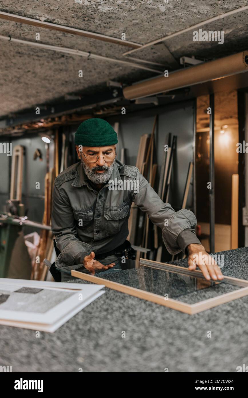 Artigiano maturo che posiziona il vetro nel telaio al banco di lavoro Foto Stock