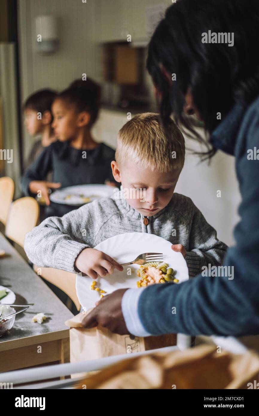 Studente maschio che getta cibo residuo in sacchetto di carta tenuto da insegnante al centro di assistenza per bambini Foto Stock