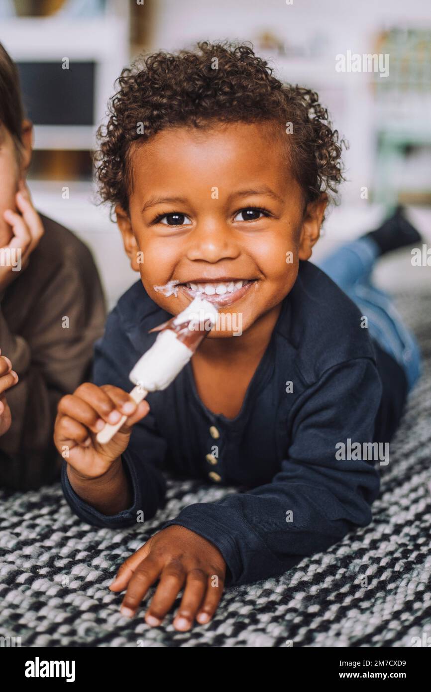 Ritratto di ragazzo felice con gelato di un compagno di classe maschile sdraiato su tappeto in asilo Foto Stock