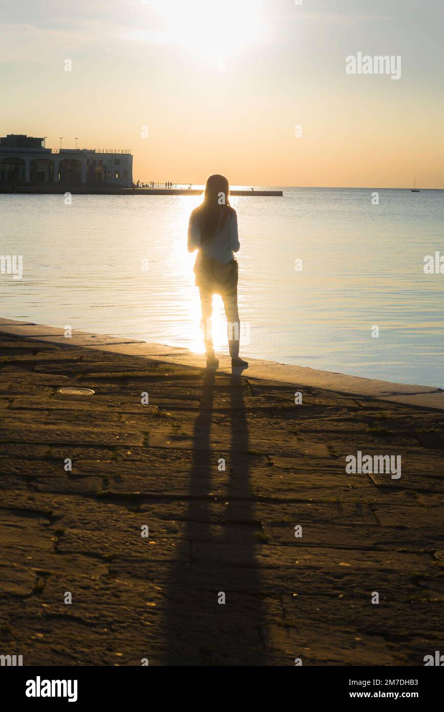 Donna da sola silhouette, vista posteriore di una giovane donna in piedi da sola alla luce del sole che guarda verso il mare. Foto Stock
