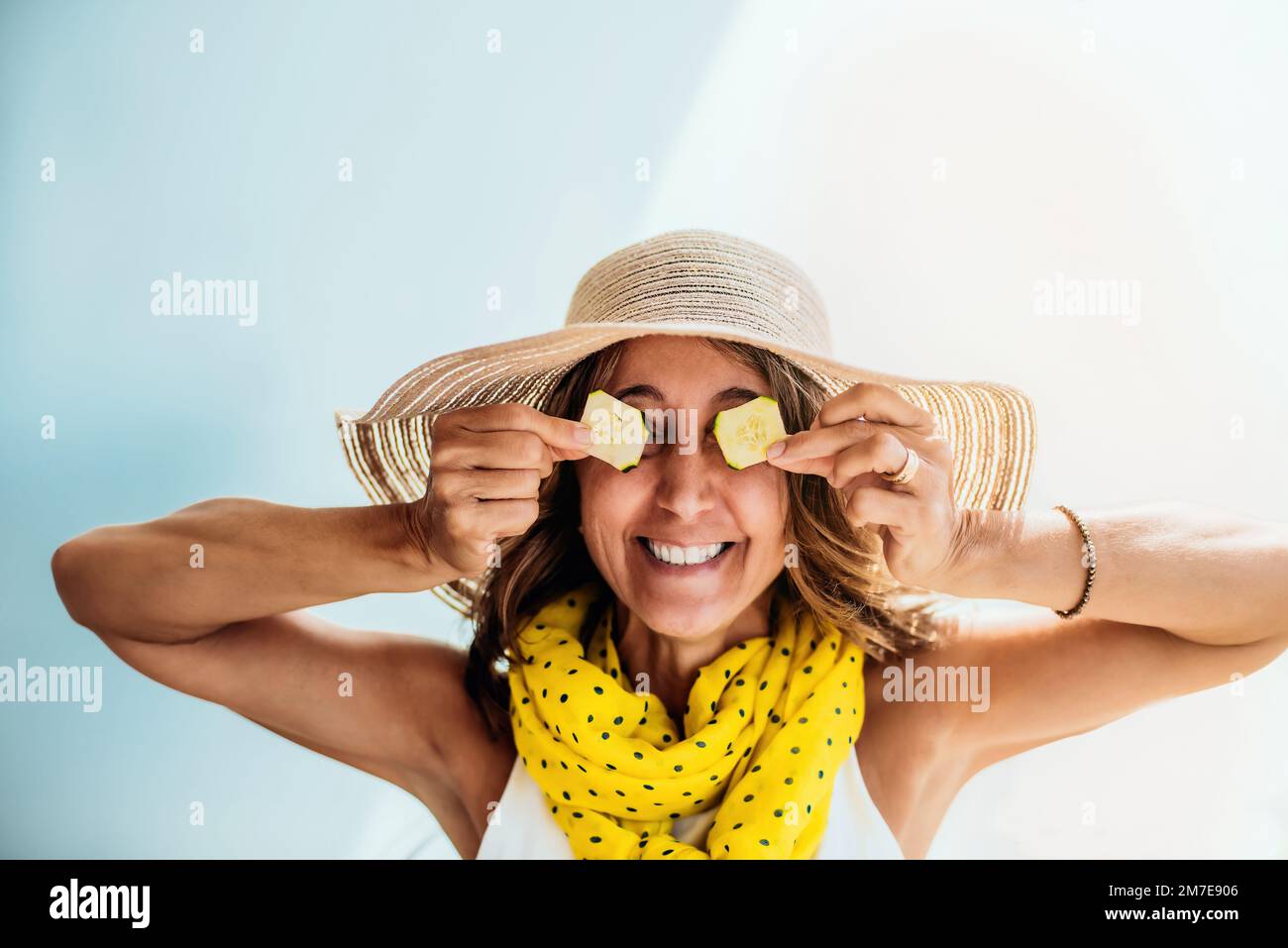 una donna di mezza età in un cappello di paglia e sciarpa gialla scherza e si diversa coprendo gli occhi con due fettine di cetriolo. concetto di salute e divertimento Foto Stock