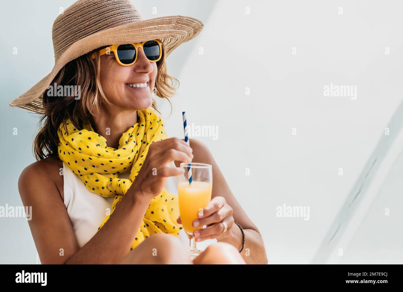 primo piano di una donna di mezza età con un cappello e bicchieri gialli che tiene in mano un succo d'arancia. concetto di vacanza e relax. Foto Stock