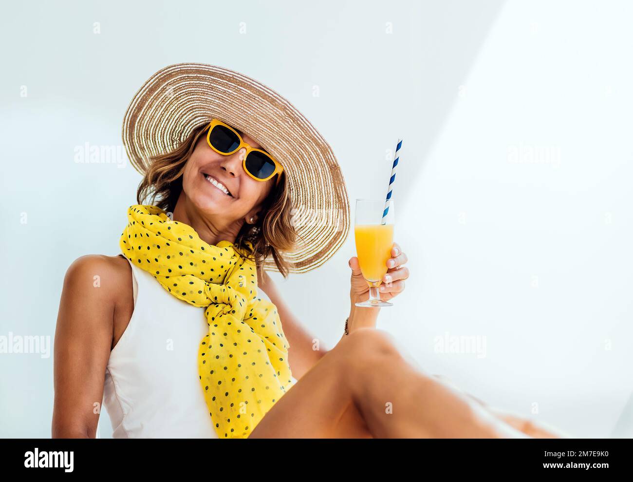 ritratto di una donna di mezza età che guarda la macchina fotografica indossando un cappello e occhiali gialli tenendo in mano un succo d'arancia. concetto di vacanza Foto Stock