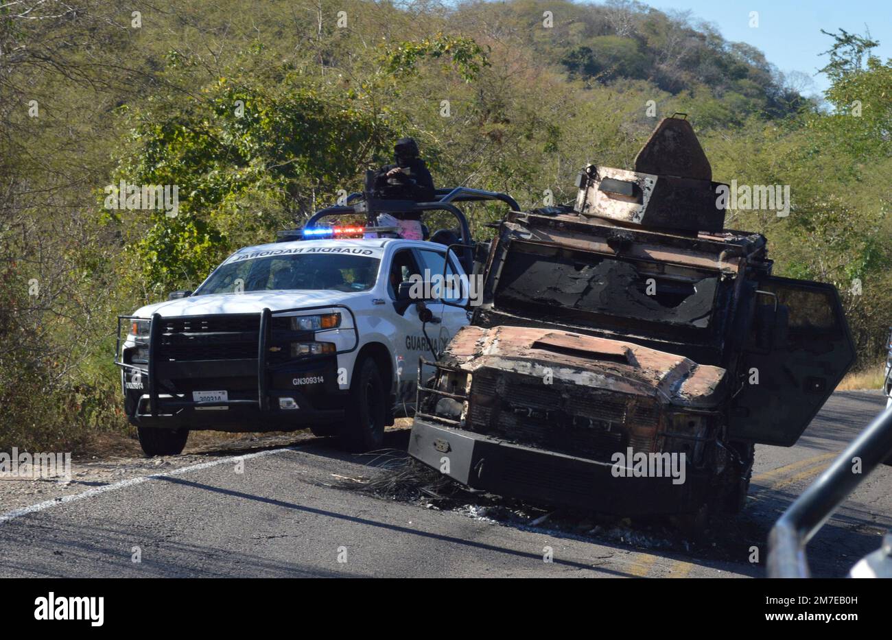 Members of the National Guard drive past a destroyed vehicle on a road near Jesus Maria, Mexico, on Saturday, Jan. 7, 2023, the small town where Ovidio Guzman was detained earlier in the week. Thursday's government operation to detain Ovidio, the son of imprisoned drug lord Joaquin "El Chapo" Guzman, unleashed firefights that killed 10 military personnel and 19 suspected members of the Sinaloa drug cartel, according to authorities. (AP Photo/Martin Urista) Foto Stock
