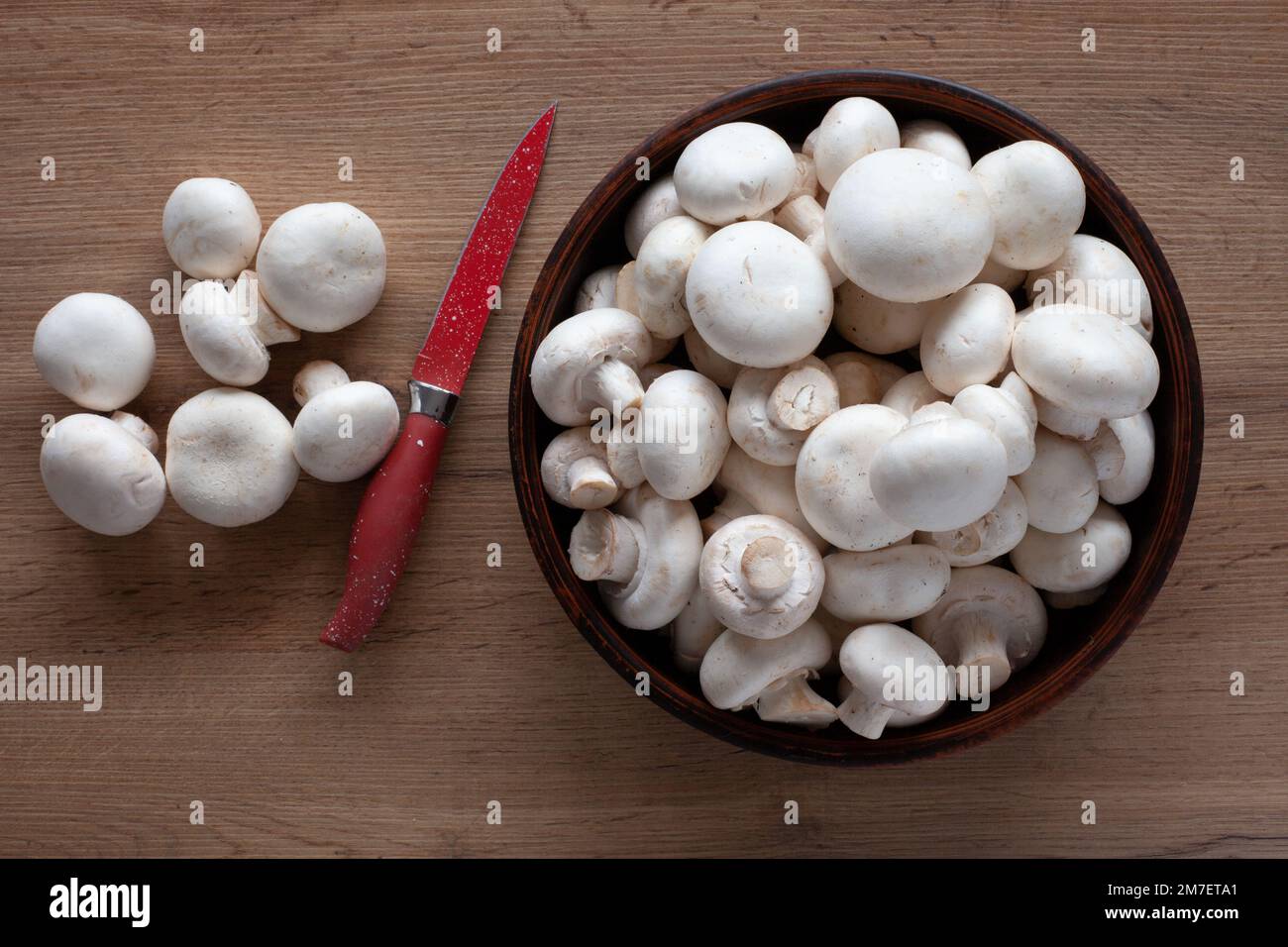 Champignons freschi sul tavolo, in una ciotola e su un asse di legno con un coltello rosso, vista dall'alto degli ingredienti vegetali per la cottura. Foto Stock