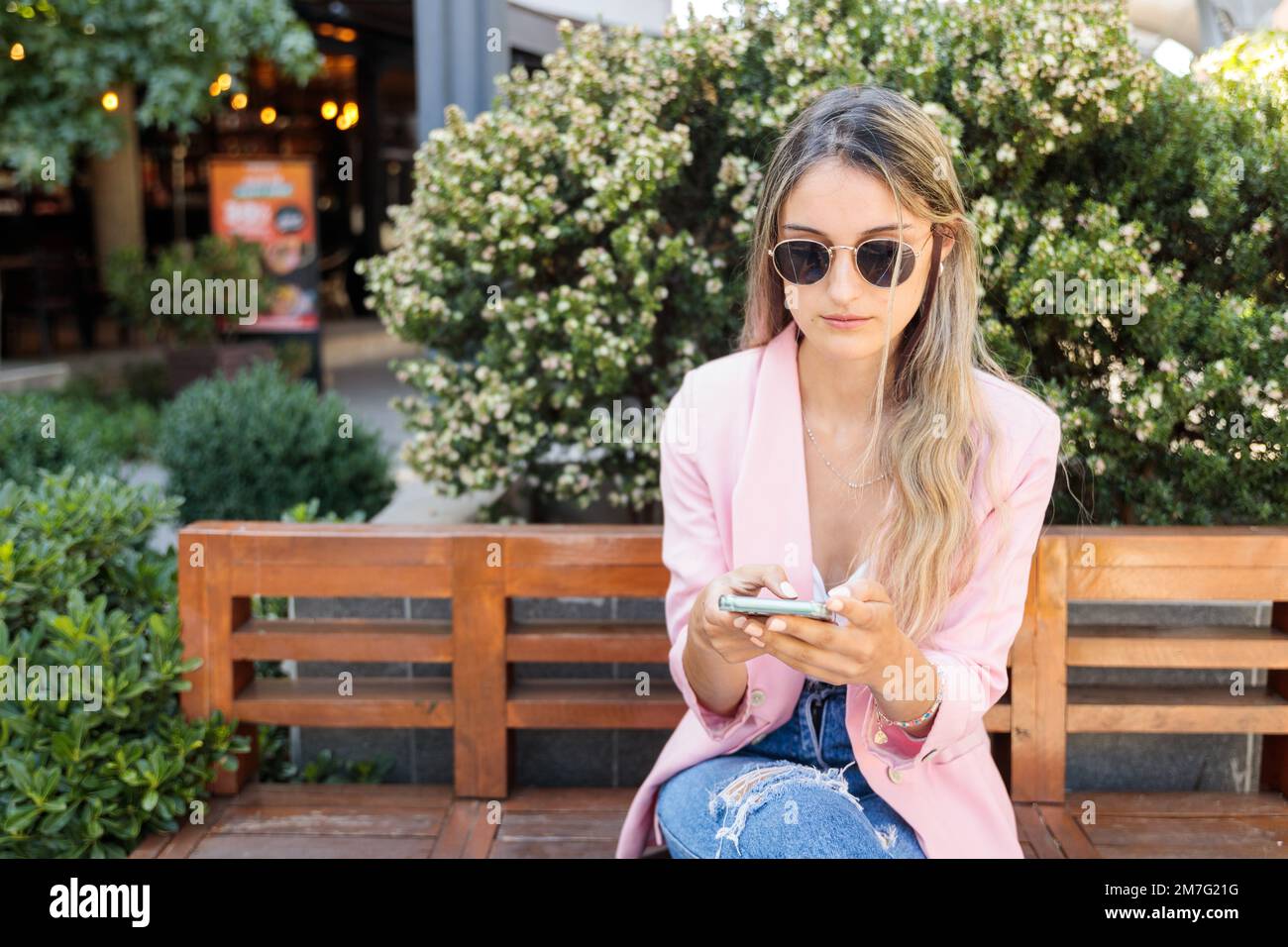 Giovane donna preoccupata, che usa lo smartphone e che siede su una panca. Dipendenza dai social media mobili Foto Stock