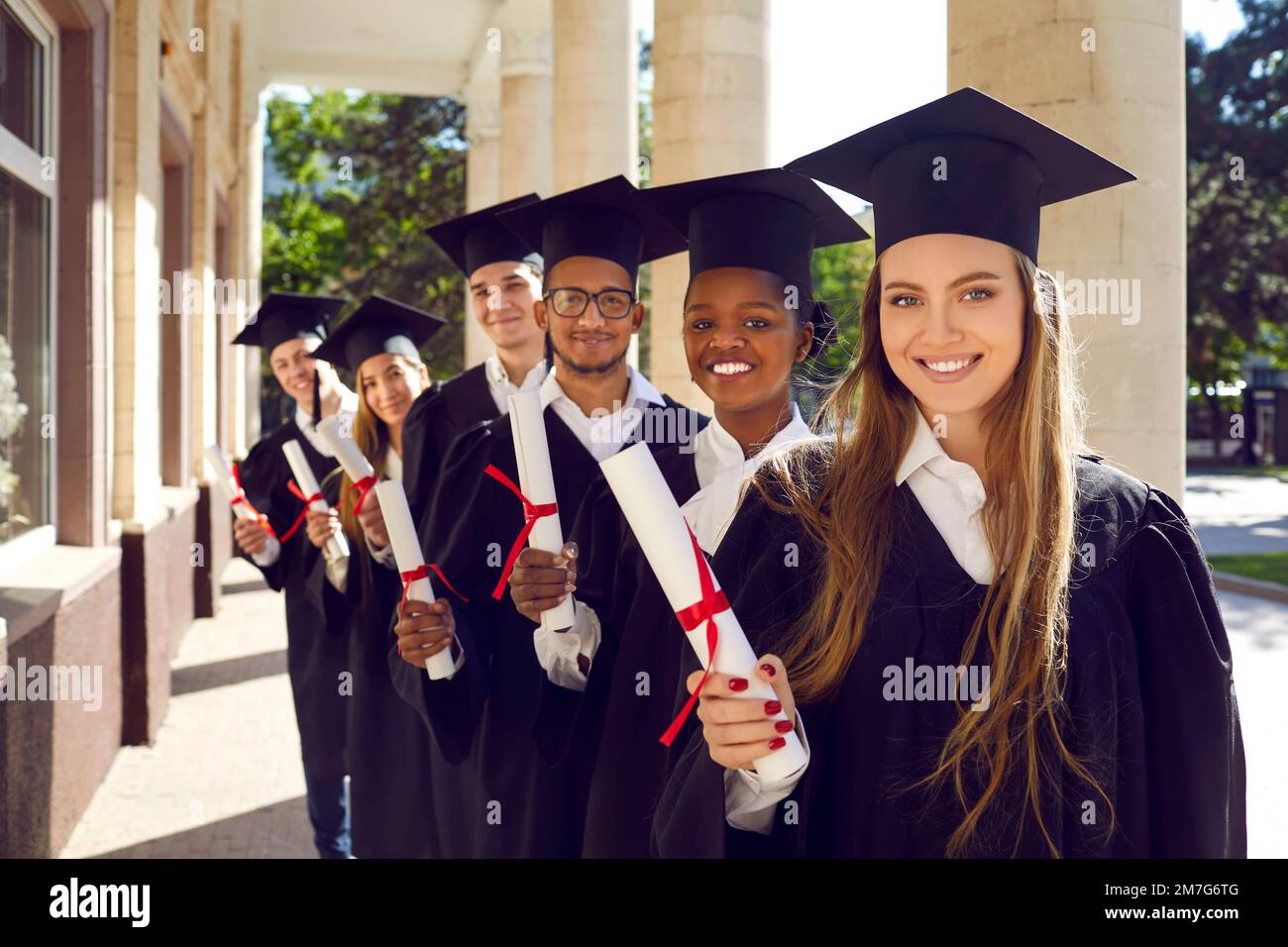 Gruppo multinazionale di allettanti studenti-laureati in tavole di mortaio cappello e abiti cerimoniali che tengono il diploma nelle loro mani. Foto Stock