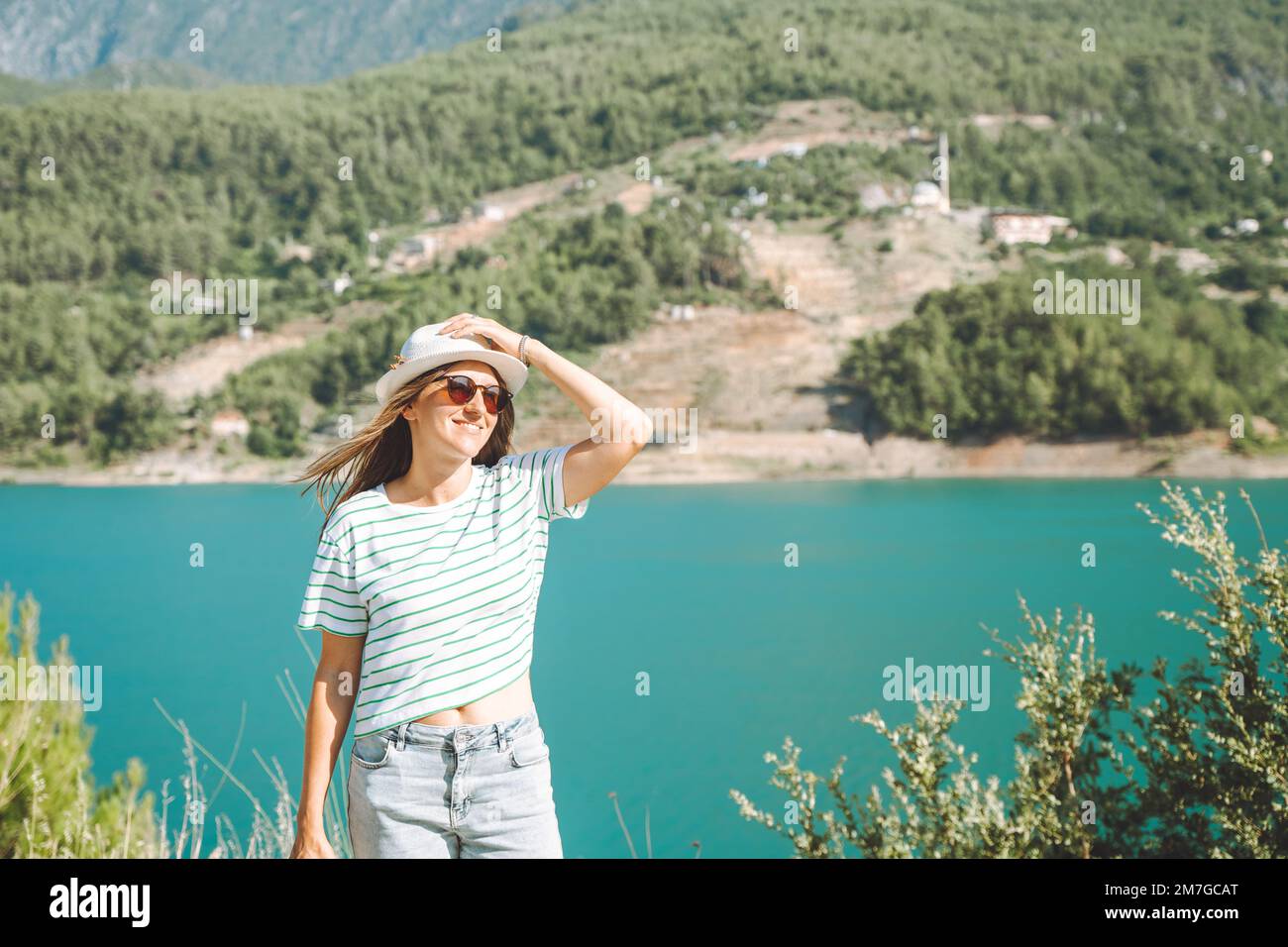 Donna sorridente in cappello e occhiali da sole con capelli selvatici in piedi vicino lago montagne sullo sfondo. Positiva giovane donna che viaggia sul lago blu all'aperto Foto Stock