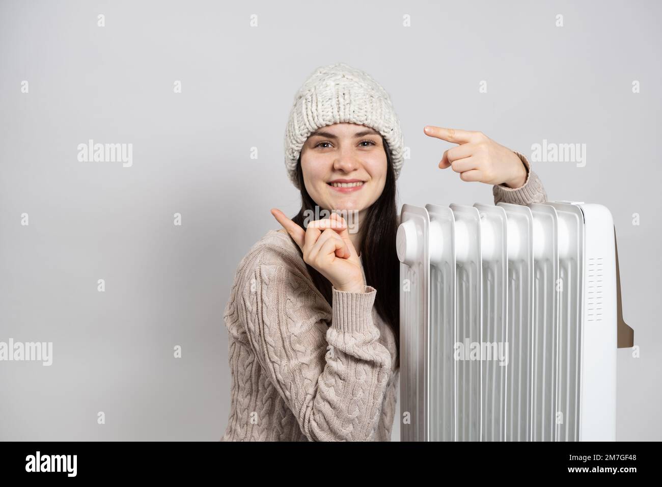 Una donna si crogiolerà vicino a un riscaldatore elettrico a olio e punta le dita verso il lato in posizione per il testo. Metodi di riscaldamento di un appartamento o di una casa Foto Stock