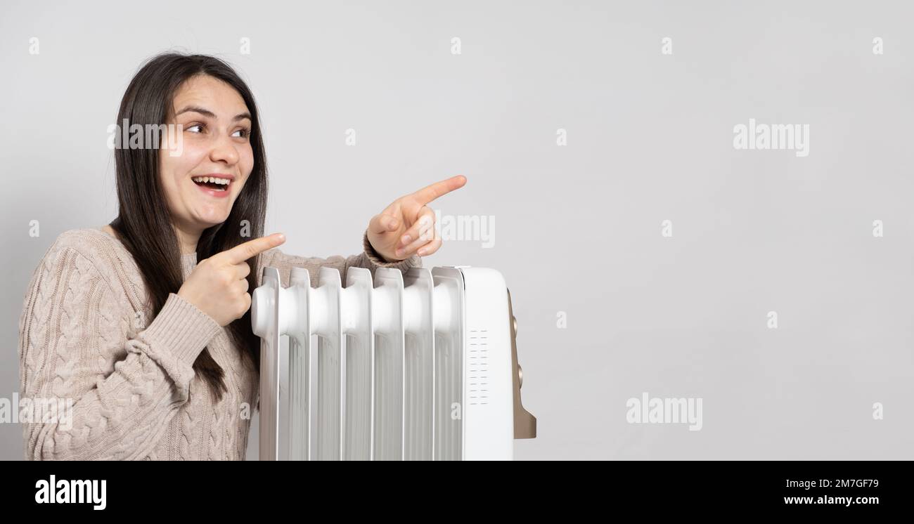 Una donna si crogiolerà vicino a un riscaldatore elettrico a olio e punta le dita verso il lato in posizione per il testo. Metodi di riscaldamento di un appartamento o di una casa Foto Stock