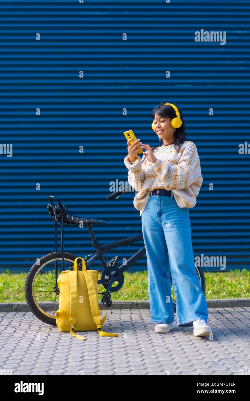 Ragazza asiatica dell'università che ascolta musica con cuffie gialle su uno sfondo blu dell'università Foto Stock