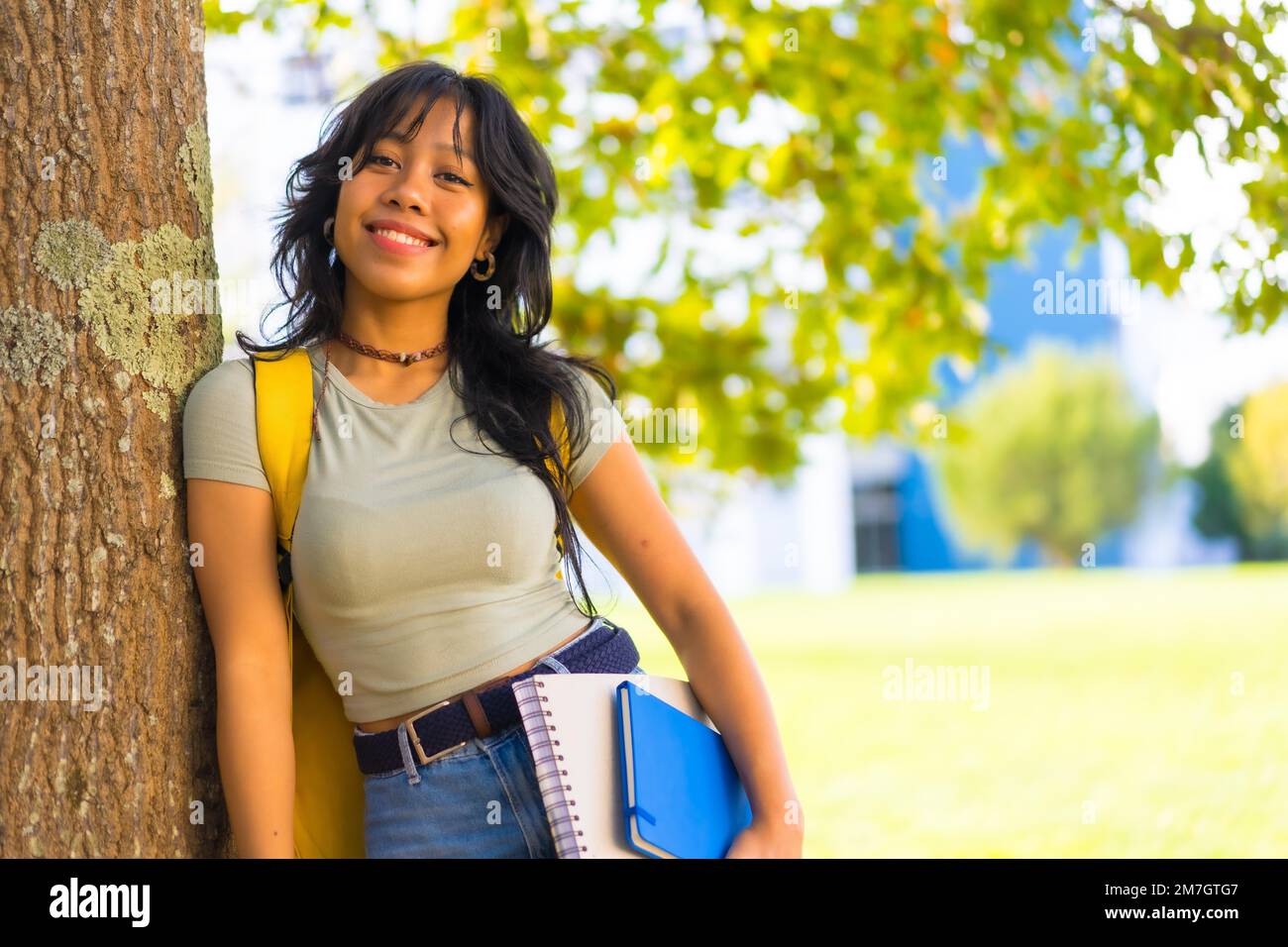 Ragazza asiatica nel campus, sorridente all'università sotto l'albero con blocco in mano e zaino Foto Stock