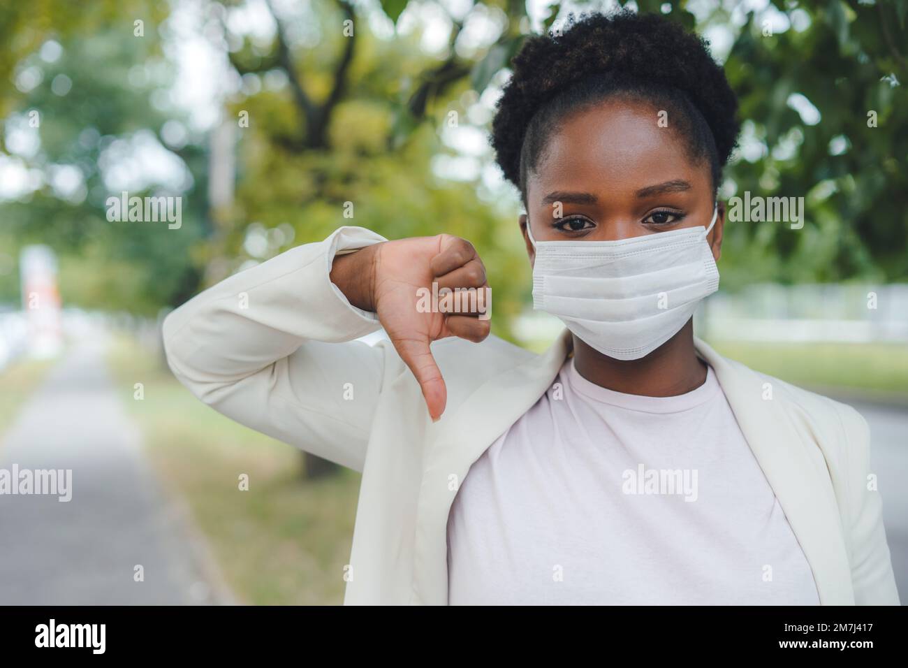 Donna d'affari africana stressata con maschera che dà pollici giù all'aperto. Misure di prevenzione dell'influenza. Concetto di assistenza sanitaria. Foto Stock