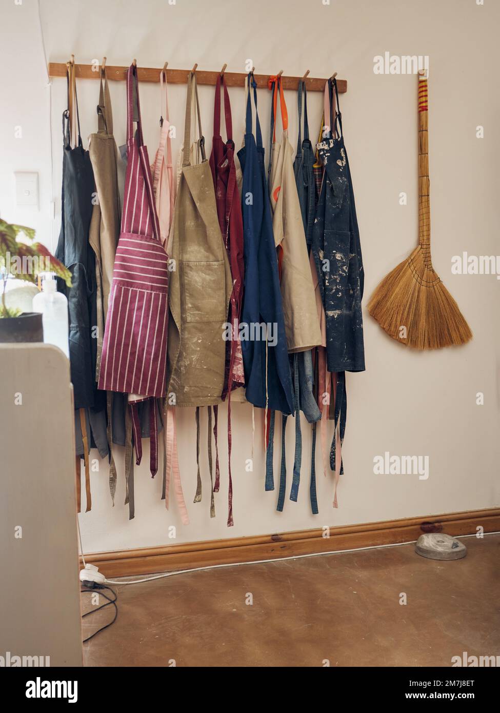 Appendiabiti, grembiule e ceramiche con una scopa in un laboratorio vuoto o studio contro il muro della vita morta per il design. Creativa, arte e abbigliamento con un Foto Stock