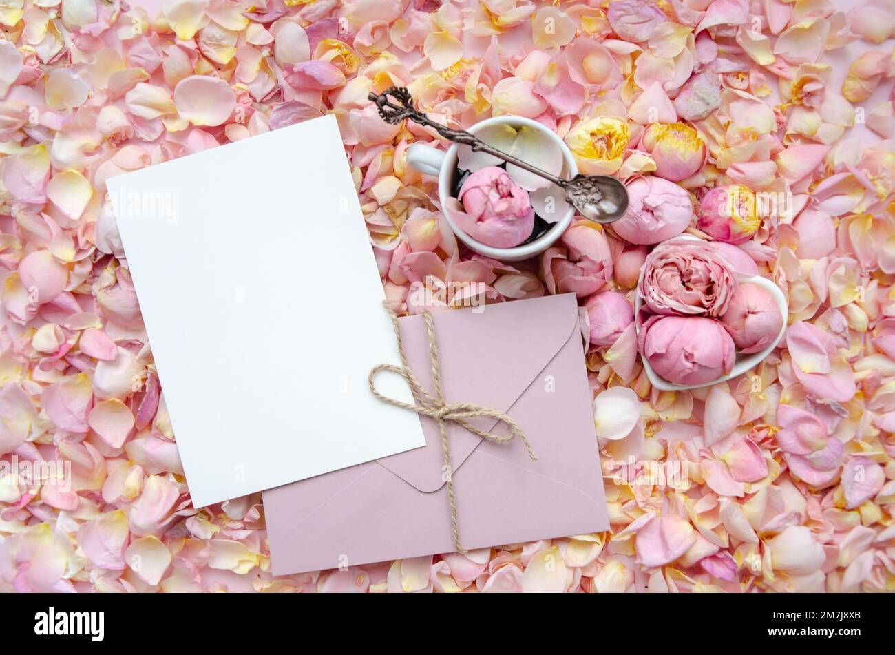 Sfondo di boccioli di rosa e petali con busta e cartoncino bianco al centro e una tazza di tè con rosa e cucchiaio da tè. Piano di giacitura, vista dall'alto. Moth Foto Stock