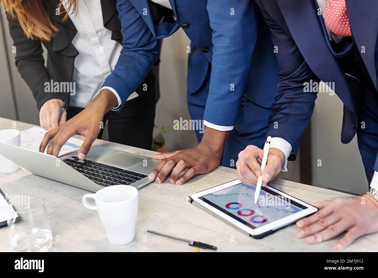 Ritagliate colleghi anonimi in un abbigliamento formale in piedi al tavolo con notebook e tablet mentre lavorate al business plan in un ufficio moderno Foto Stock