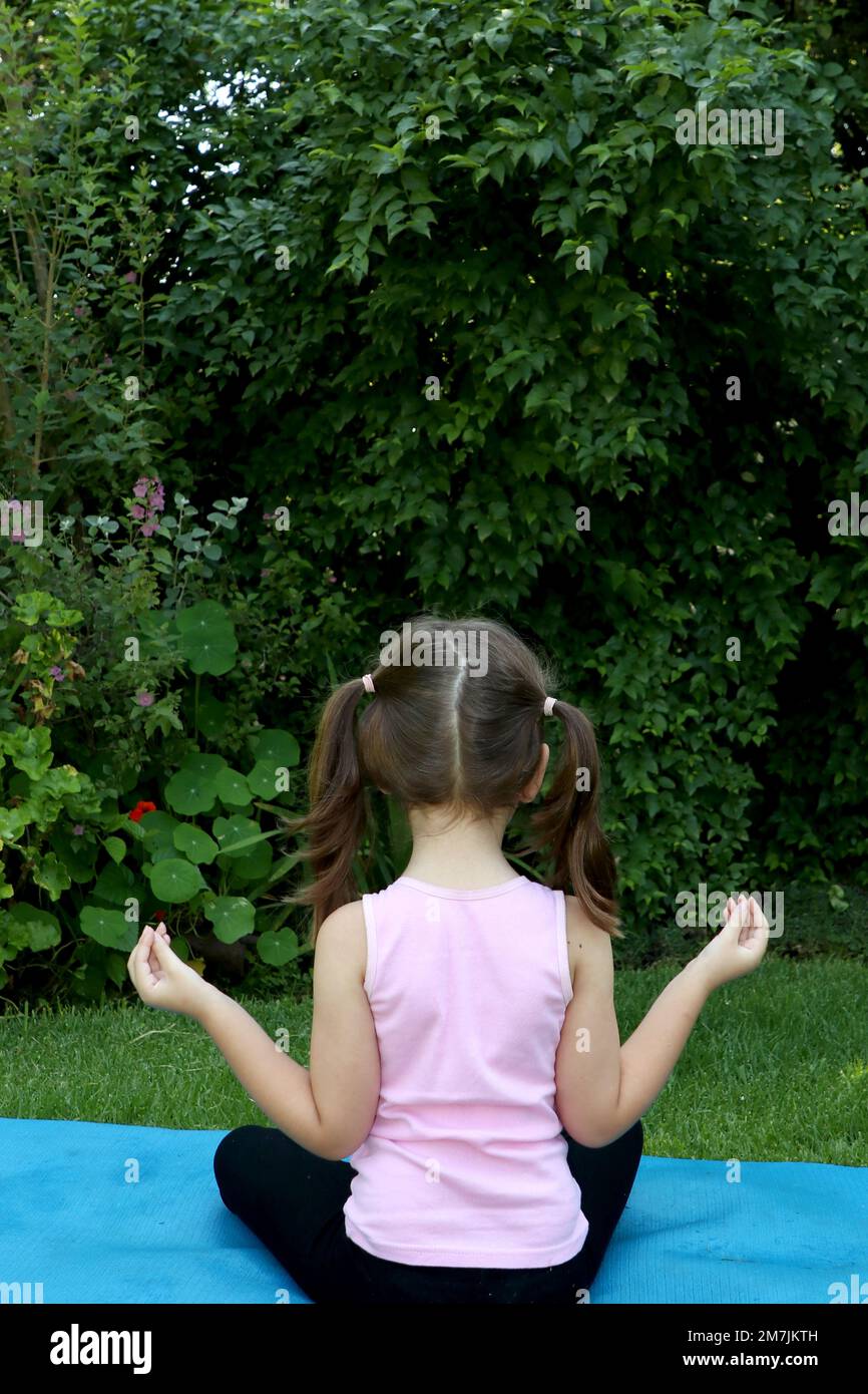 Ragazza giovane di 7-10 anni che fa yoga posa in giardino con pigtail su un tappeto di yoga blu Foto Stock