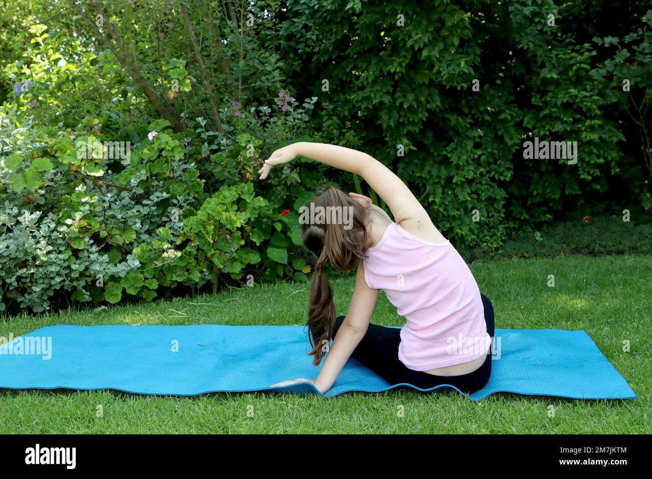 Ragazza giovane di 7-10 anni che fa yoga posa in giardino con pigtail su un tappeto di yoga blu Foto Stock