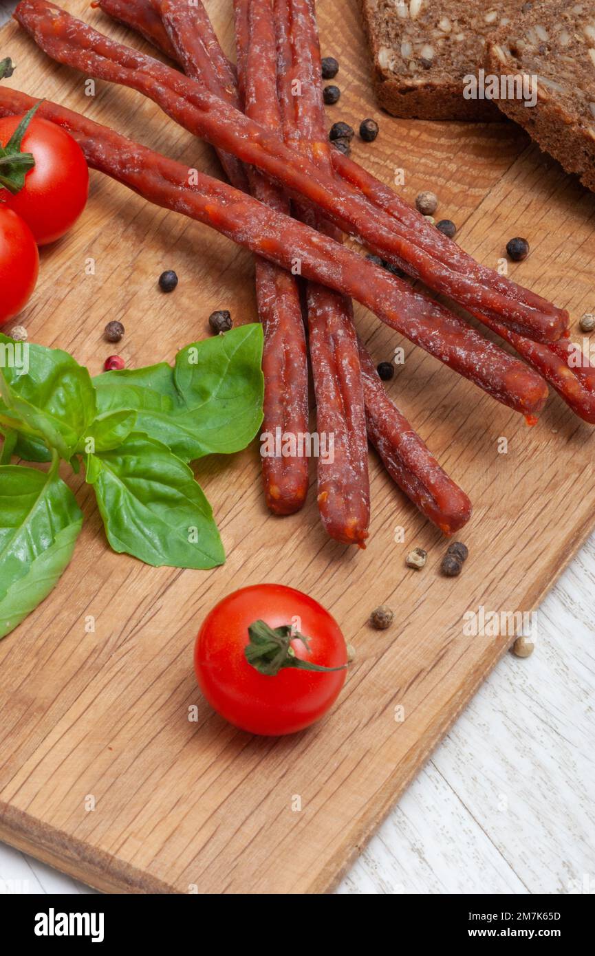 Sottili salsicce da caccia su un asse di legno decorato con pomodori e basilico Foto Stock