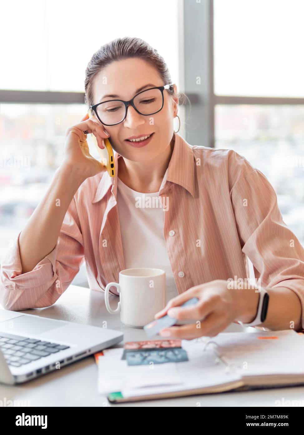 Donna lavora con computer portatile, carte di credito e blocco note. Donna sorridente sta controllando le carte di credito o la carta d'identità. Ufficio moderno con finestre panoramiche. Foto Stock