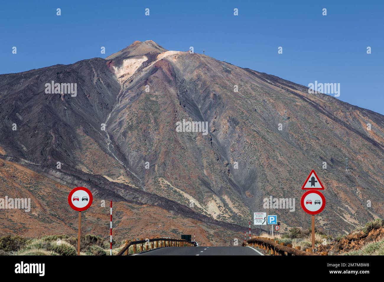 Massiccio vulcano con pendici arancioni e verdi nere e tenui viste dall'autostrada vuota sottostante, con segnaletica stradale in primo piano, sotto Foto Stock