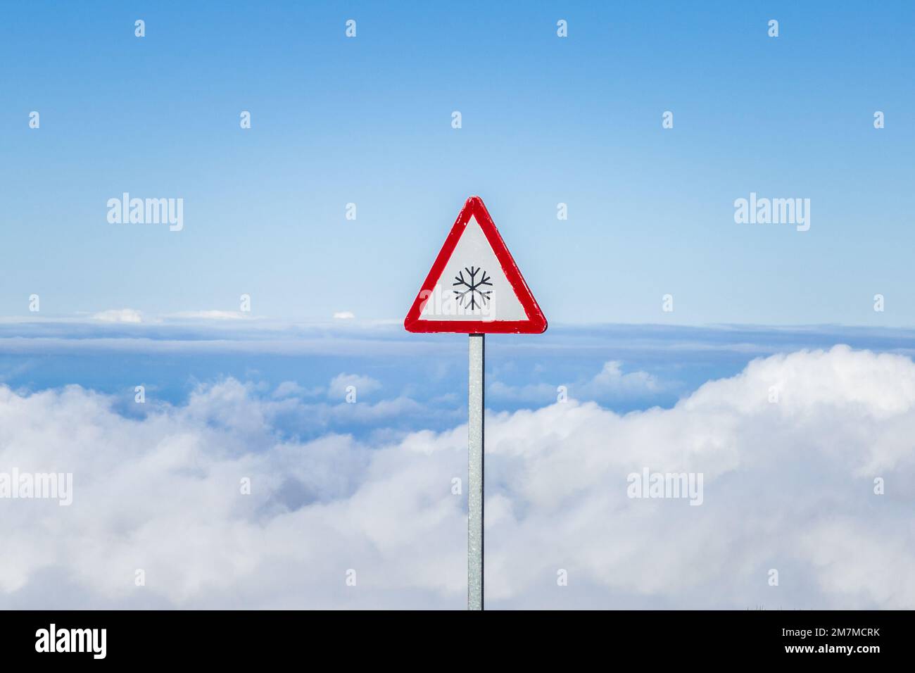 Cartello stradale triangolare per la neve sulla strada, che si affaccia su uno spesso strato di nuvole bianche sopra l'oceano e l'isola, sotto un cielo blu Foto Stock