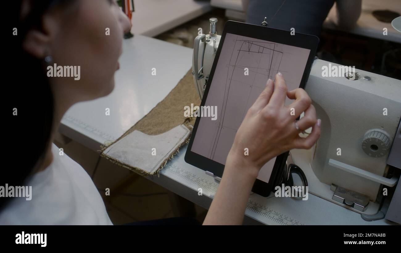 Il sarto femminile si siede sulla macchina da cucire con tablet digitale e zoom dell'immagine. Orologi da parrucchiere o da cucire taglie di abbigliamento personalizzato a bozzetto e tuta da cucire. Laboratorio Atelier. Concetto di moda. Foto Stock