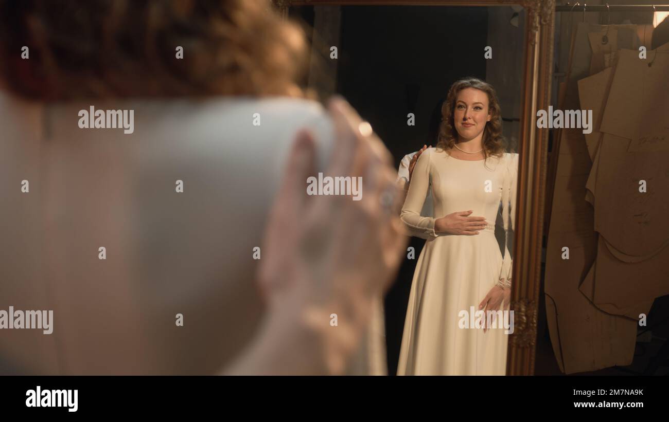 La donna sartoriale si accartoria sul vestito da sposa davanti allo specchio. Sposa felice sul vestito da sposa adatto in atelier designer di lusso o salone di nozze. Concetto di moda, fatto a mano e couturier. Foto Stock