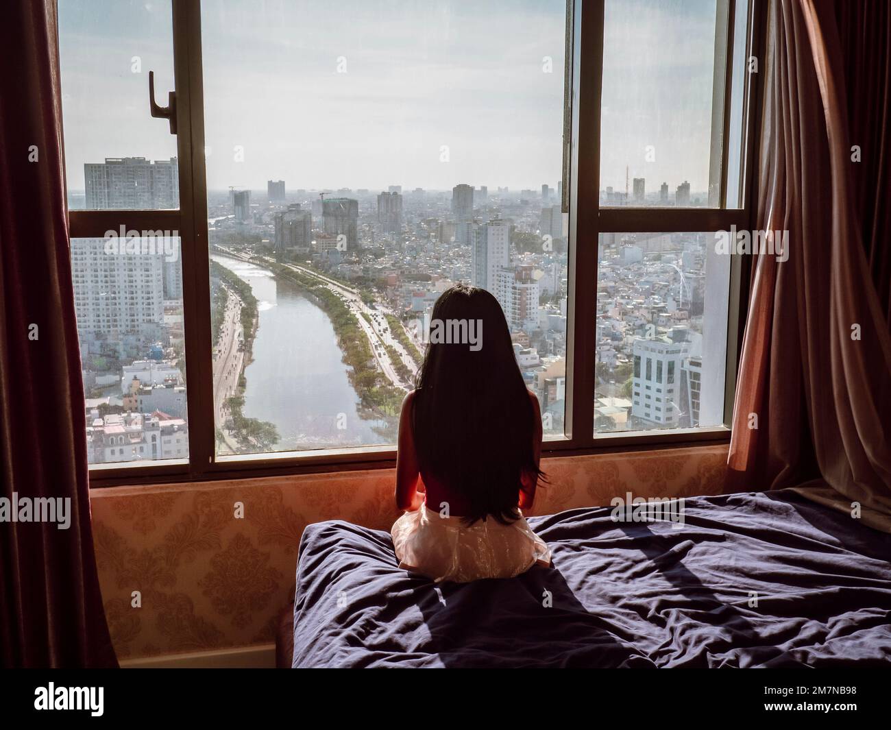 Vista posteriore di una donna asiatica seduta su un letto e guardando attraverso la finestra verso la città di ho Chi Minh, Vietnam Foto Stock