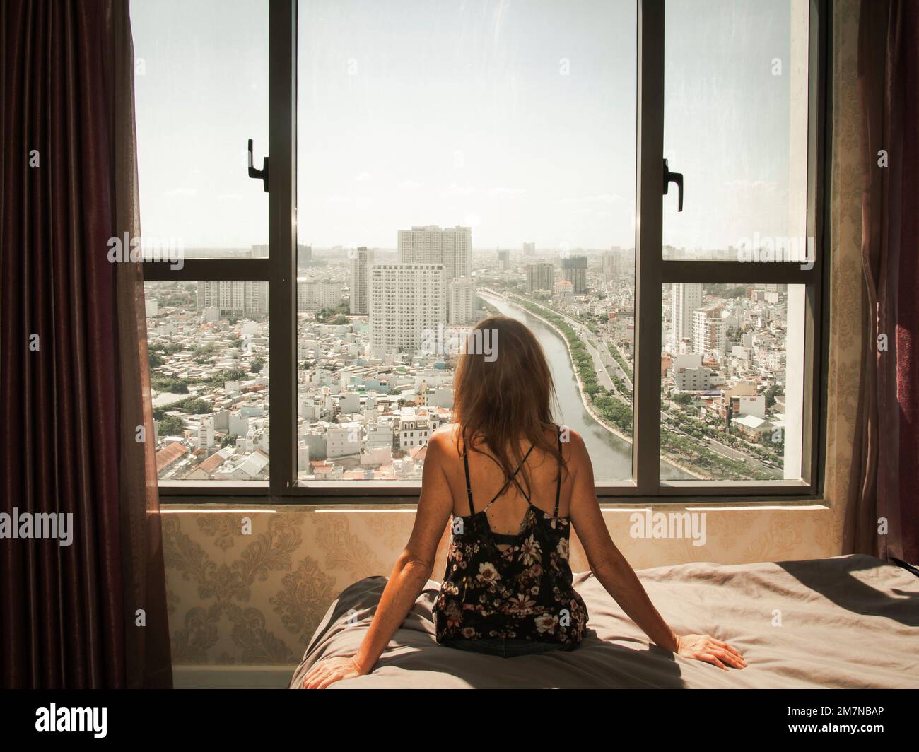 Vista posteriore di una donna caucasica seduta su un letto e guardando attraverso la finestra verso la città di ho Chi Minh, Vietnam - concetto di isolamento casa Foto Stock