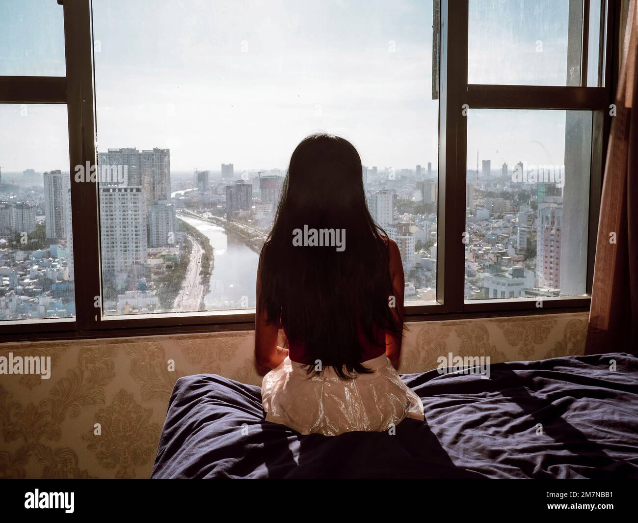 Vista posteriore di una donna asiatica seduta su un letto e guardando attraverso la finestra verso la città di ho Chi Minh, Vietnam Foto Stock