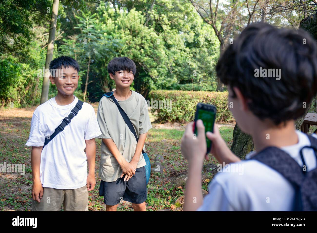 Un ragazzo con un telefono cellulare che fotografa due ragazzi di 13 anni fianco a fianco, all'aperto in un parco d'estate. Foto Stock