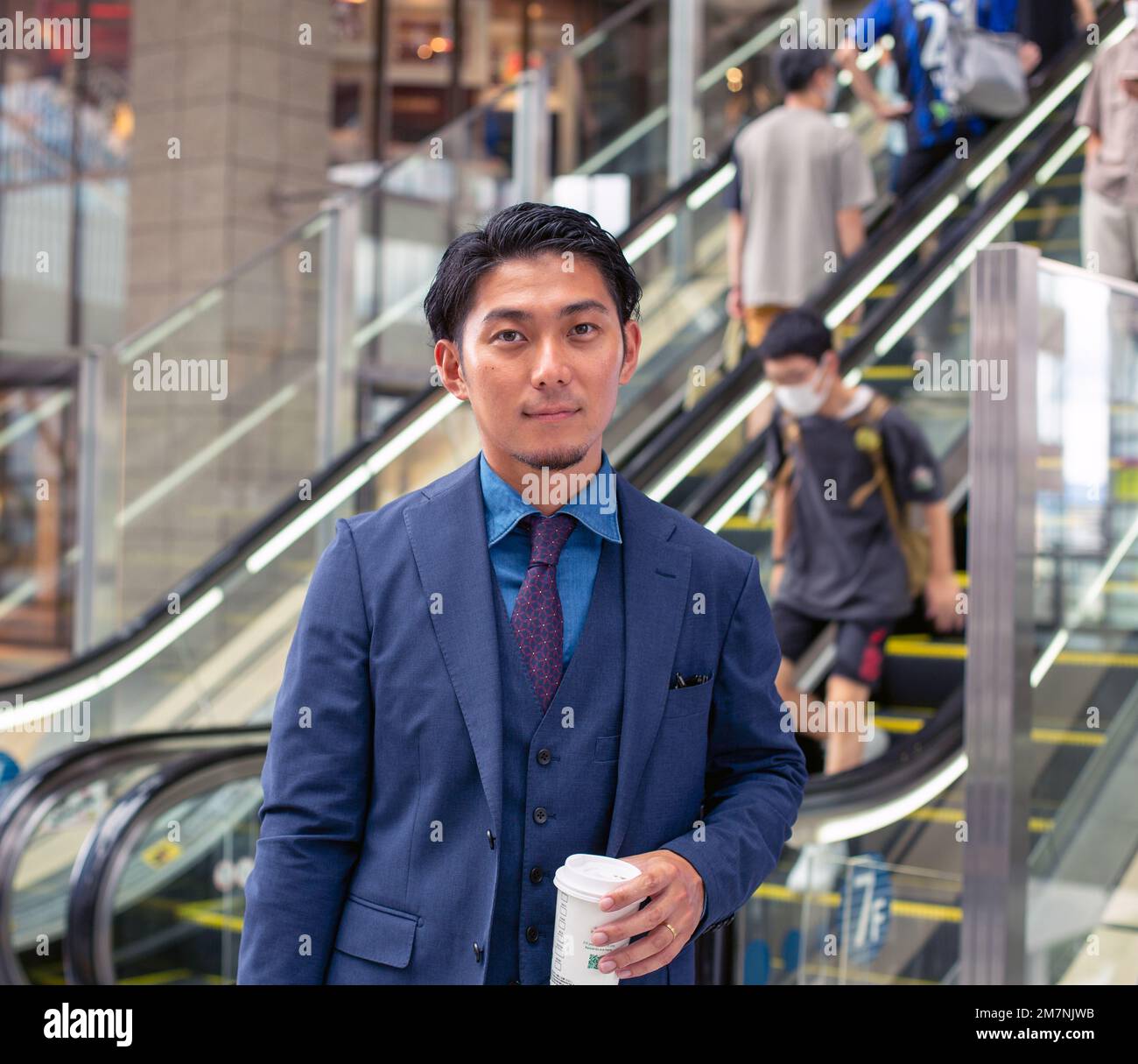 Un giovane uomo d'affari in tuta blu in movimento in una zona del centro città, con una valigetta e una tazza di caffè. Foto Stock