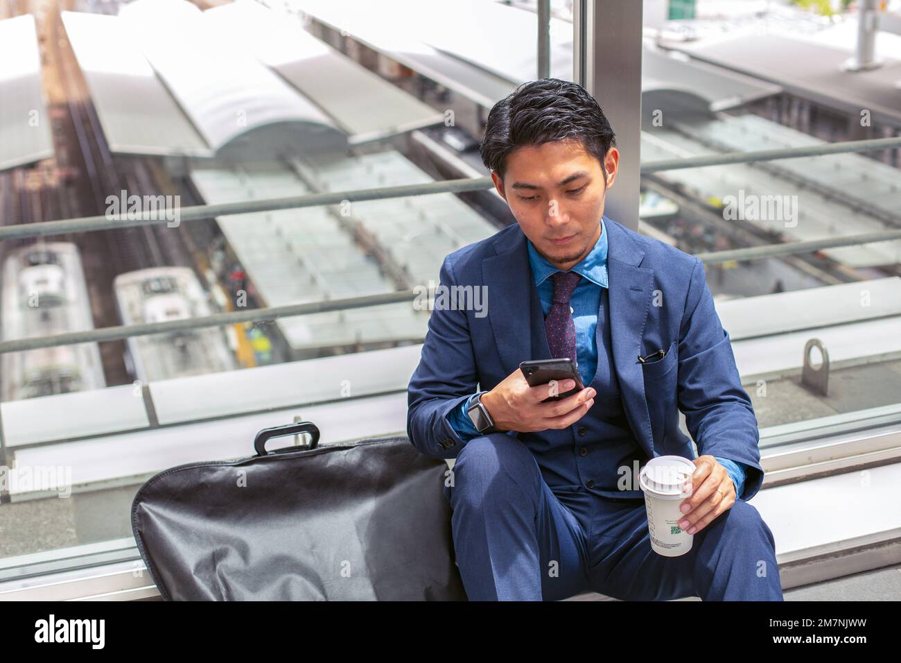 Un giovane uomo d'affari in una tuta blu in una città, guardando lo schermo del suo telefono cellulare, testando o leggendo un messaggio. Foto Stock