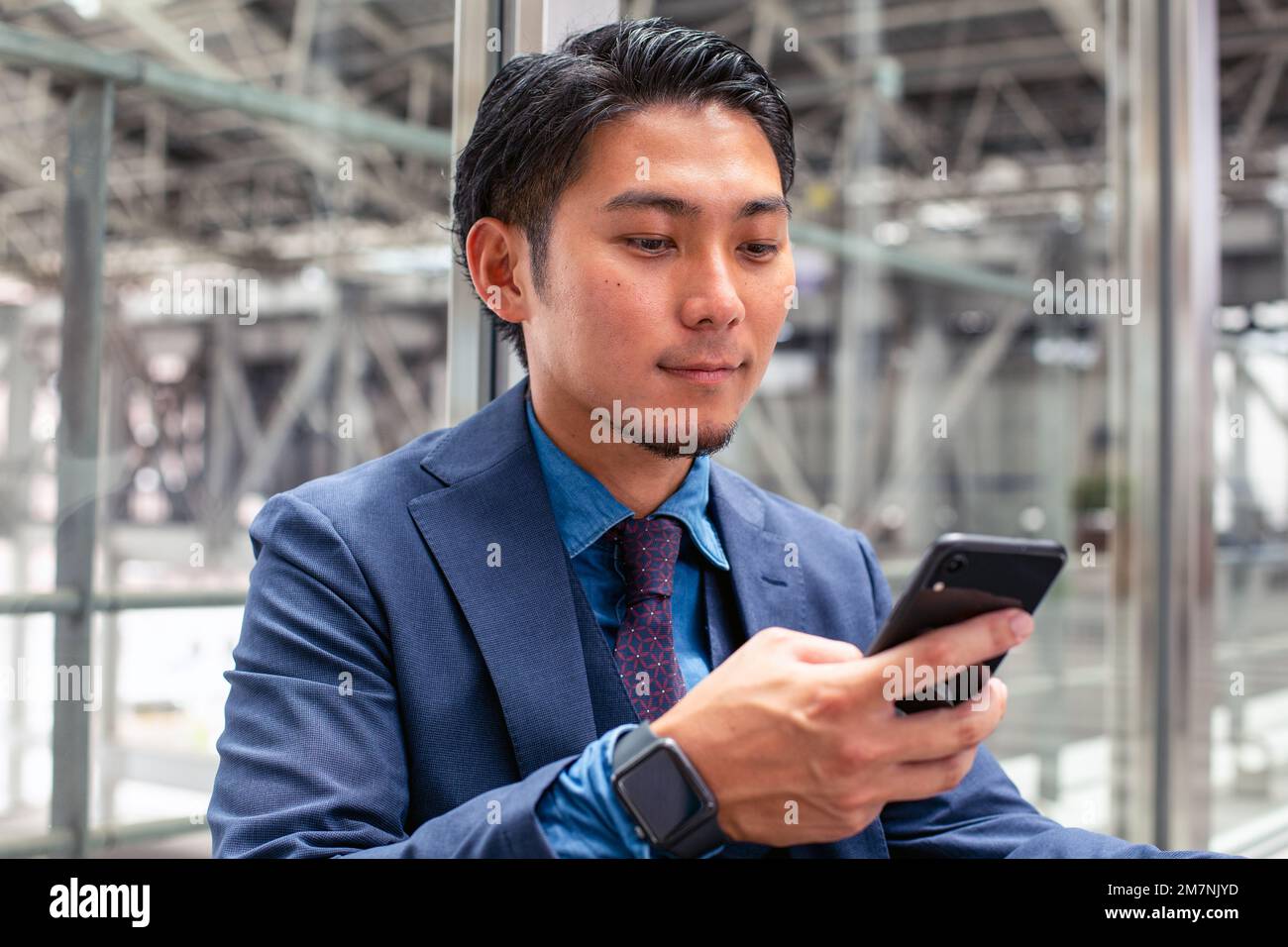 Un giovane uomo d'affari in una tuta blu in una città, guardando lo schermo del suo telefono cellulare, testando o leggendo un messaggio. Foto Stock
