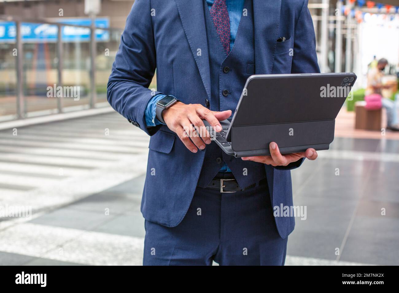 Un giovane uomo d'affari in tuta blu in movimento in una zona del centro città, in piedi e utilizzando il suo tablet digitale. Foto Stock