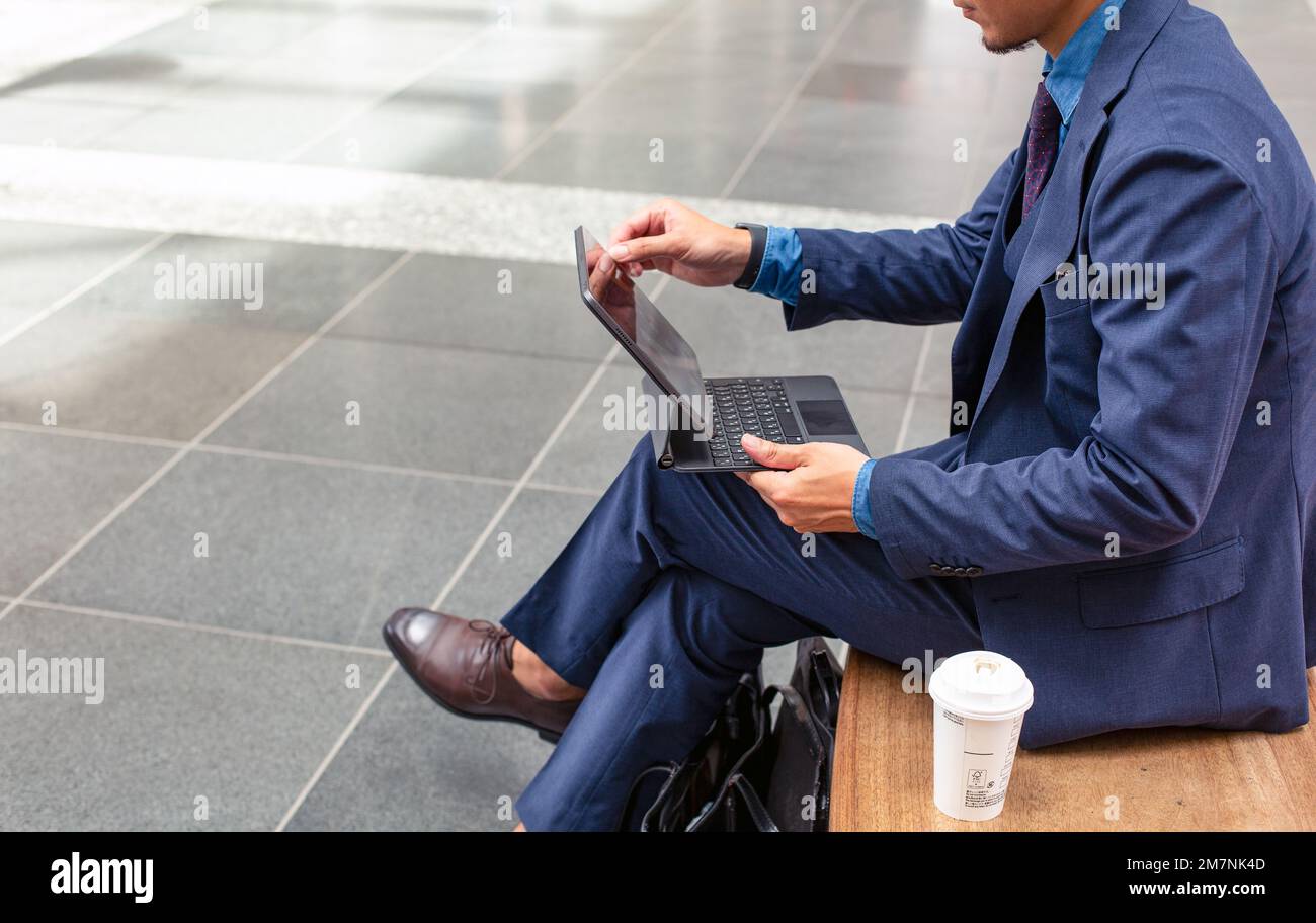 Un giovane uomo d'affari in tuta blu in movimento in una zona del centro città, seduto su una panca utilizzando un tablet digitale con schermo a sfioramento. Foto Stock