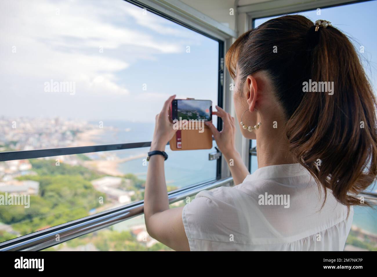 Una donna giapponese matura che utilizza il suo telefono cellulare per scattare foto da una cabina della funivia della città e del paesaggio sottostante. Foto Stock