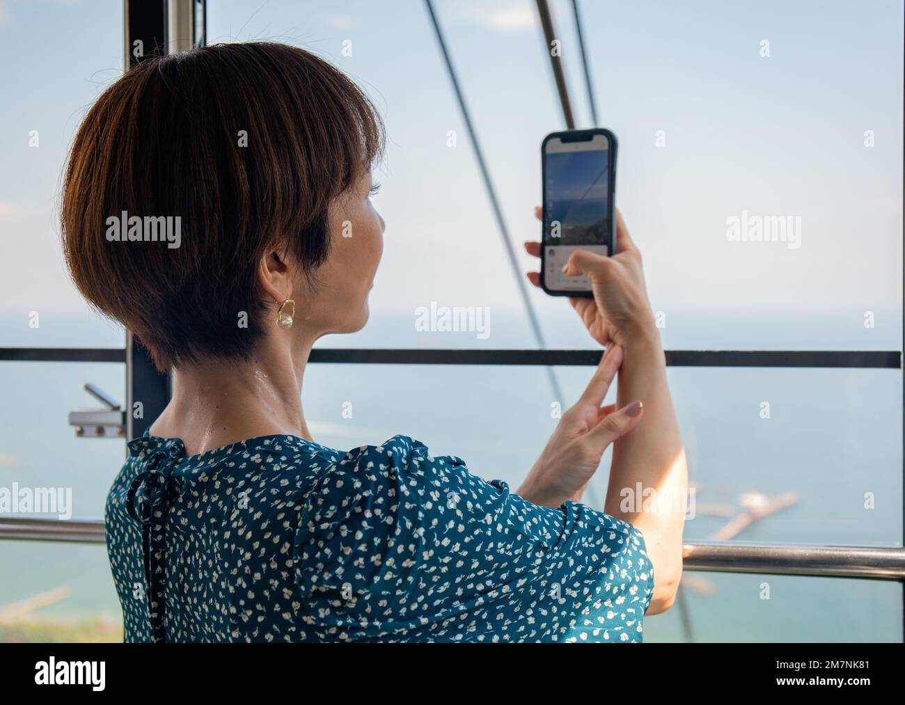 Una donna giapponese matura che utilizza il suo telefono cellulare per scattare foto da una cabina della funivia della città e del paesaggio sottostante. Foto Stock