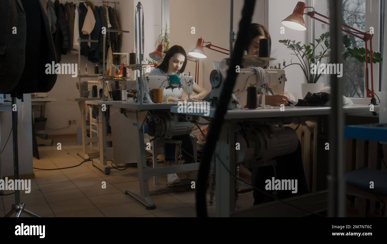 Marinaio femminile seduto a macchine da cucire e lavorando su abbigliamento personalizzato. Misura nastri e computer portatile con uno schizzo di abito da sposa al tavolo. Atelier o laboratorio di cucito. Concetto di moda. Foto Stock