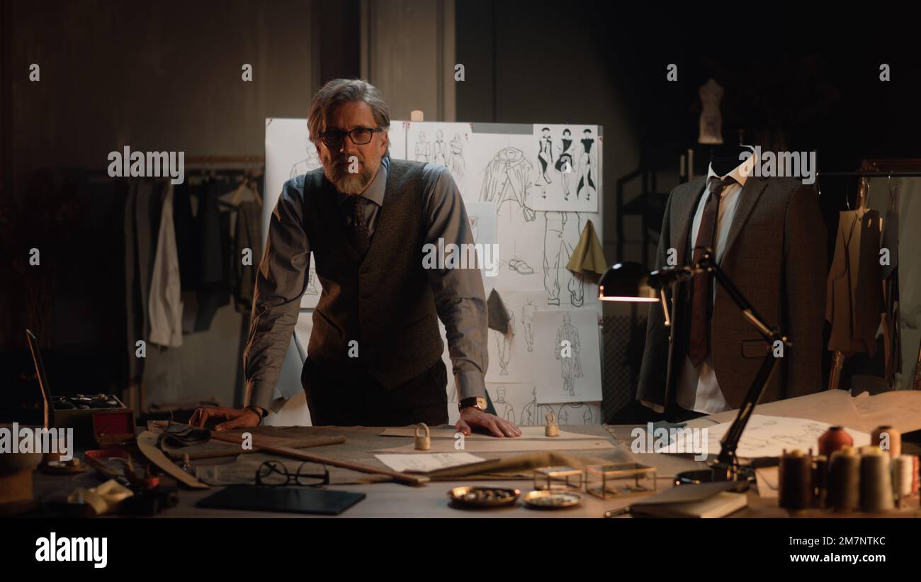 Uomo maturo sartoriale lavori in lussuoso designer atelier o studio di sartoria, guarda la fotocamera. Tavolo con fili, tessuto e utensili. Abiti e tavola su misura con schizzi sullo sfondo. Concetto di moda. Foto Stock