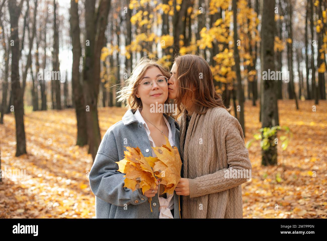 Madre bacia in guancia figlia adolescente in foresta soleggiata. Le ragazze tengono caduti fogli di acero giallo e camminano nel parco autunnale. Foto Stock